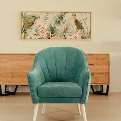Cadeira verde água estofada com pernas brancas numa sala com móvel de madeira e quadro de pássaros