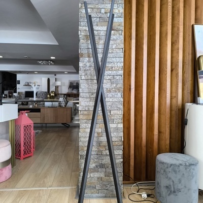 Cabideiro metálico preto em formato de X junto a parede de pedra e madeira, ao lado de tamborete cinza e candeeiro branco.