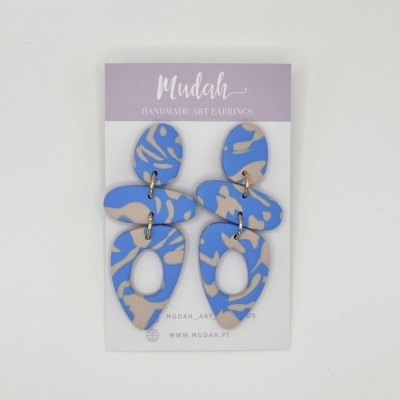 Brincos artesanais azul e bege com padrão abstrato num cartão de apresentação lilás e branco