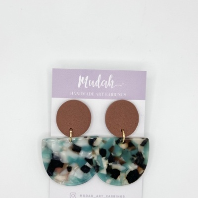 Brincos artesanais com parte superior castanha redonda e parte inferior semicircular marmoreada em azul, branco, preto e castanho, em cartão da marca Mudah.