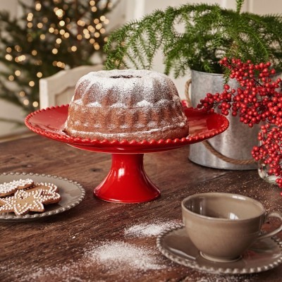 Bolo bundt com açúcar em pó numa base vermelha, chávena e biscoitos, com decoração natalícia ao fundo