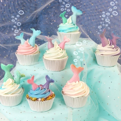 Cupcakes decorados com caudas de sereia em tons pastel, sobre tecido azul claro e fundo azul com bolhas.