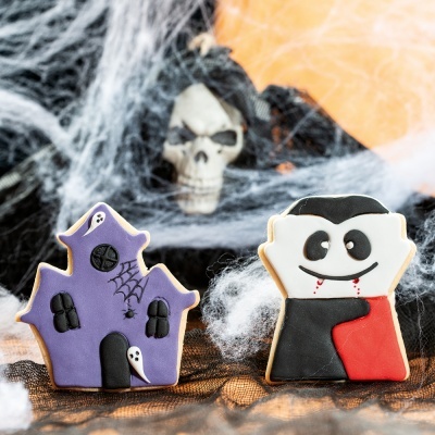 Biscoitos decorados de Halloween em forma de casa assombrada e vampiro com decoração assustadora ao fundo.
