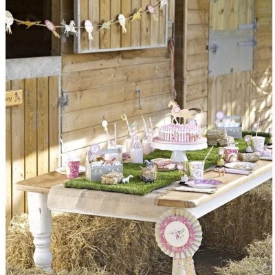 Mesa de festa infantil decorada com bolo e utensílios cor-de-rosa num celeiro de madeira com feno no chão