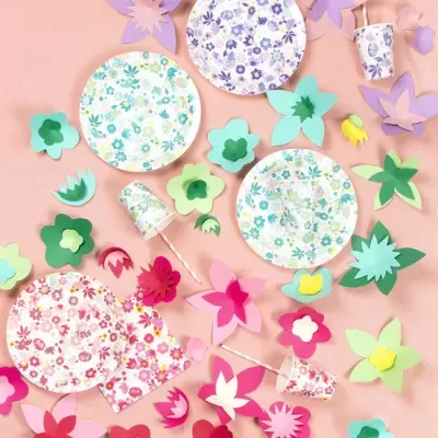 Conjunto de louça descartável floral com pratos, copos e guardanapos em tons pastel, decorados com flores, sobre superfície rosa com flores de papel coloridas