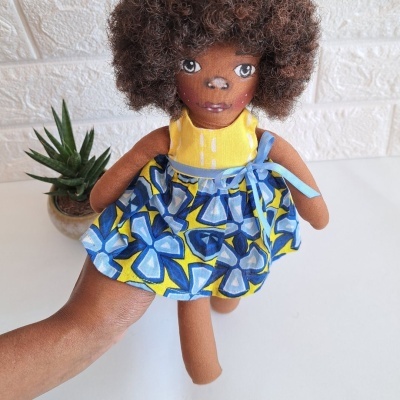 Boneca de pano com vestido amarelo e azul, com cabelo encaracolado e planta ao lado.