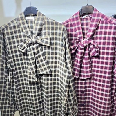 Duas camisas xadrez com laço no pescoço, uma cinza e bege e outra rosa e branco, penduradas em cabides pretos.