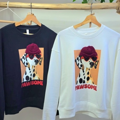 Sweatshirts preta e branca com estampa de dálmata usando chapéu roxo e óculos vermelhos, texto PAWSOME em vermelho, penduradas em cabides de madeira