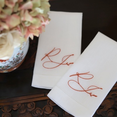 Toalhas de tecido branco bordadas com letras laranja sobre mesa de madeira com vaso floral