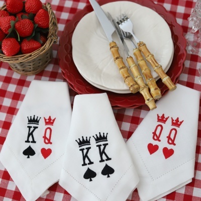 Guardanapos brancos com bordados de cartas K e Q em preto e vermelho numa mesa com toalha xadrez vermelha e branca