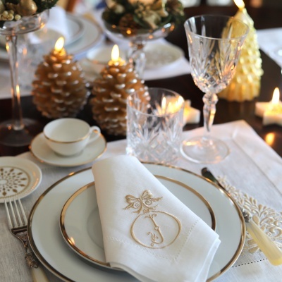 Mesa posta com pratos brancos com borda dourada, guardanapo bordado, copos de vidro lapidado e velas decorativas douradas