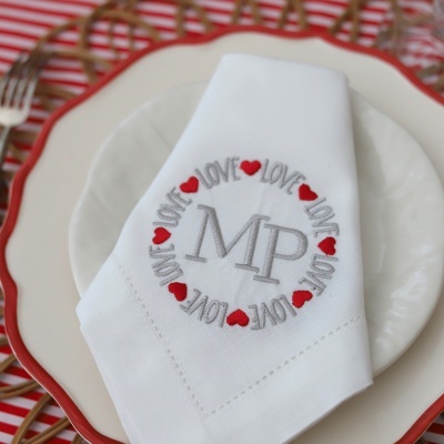 Guardanapo branco bordado com as letras MP e motivos de corações vermelhos e palavra LOVE em círculo, sobre pratos brancos em mesa decorada.