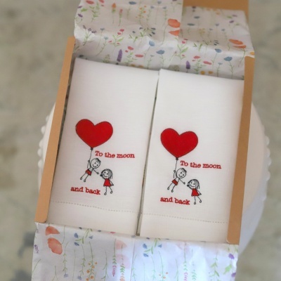 Dois panos de cozinha brancos com desenho de crianças e balão vermelho com texto To the moon and back em embalagem floral.