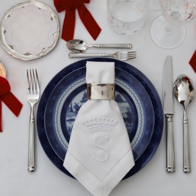 Mesa posta com pratos azuis e brancos, guardanapo bordado branco com argola prateada, talheres prateados e copos transparentes