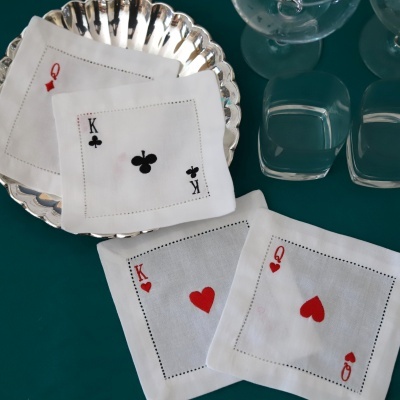 Guardanapos de tecido brancos bordados com símbolos de cartas de baralho ao centro, sobre mesa verde com prato metálico e copos claros.