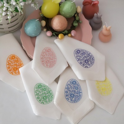 Guardanapos de tecido branco com desenhos de ovos florais coloridos sobre mesa com velas em forma de ovo e coelho e prato rosa.