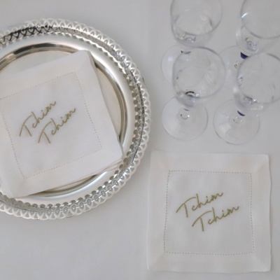 Guardanapos brancos com bordado dourado Tchim Tchim, pratos prateados e copos de vidro numa mesa branca