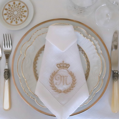 Conjunto de pratos com borda dourada, guardanapo branco bordado, talheres e copos num fundo branco