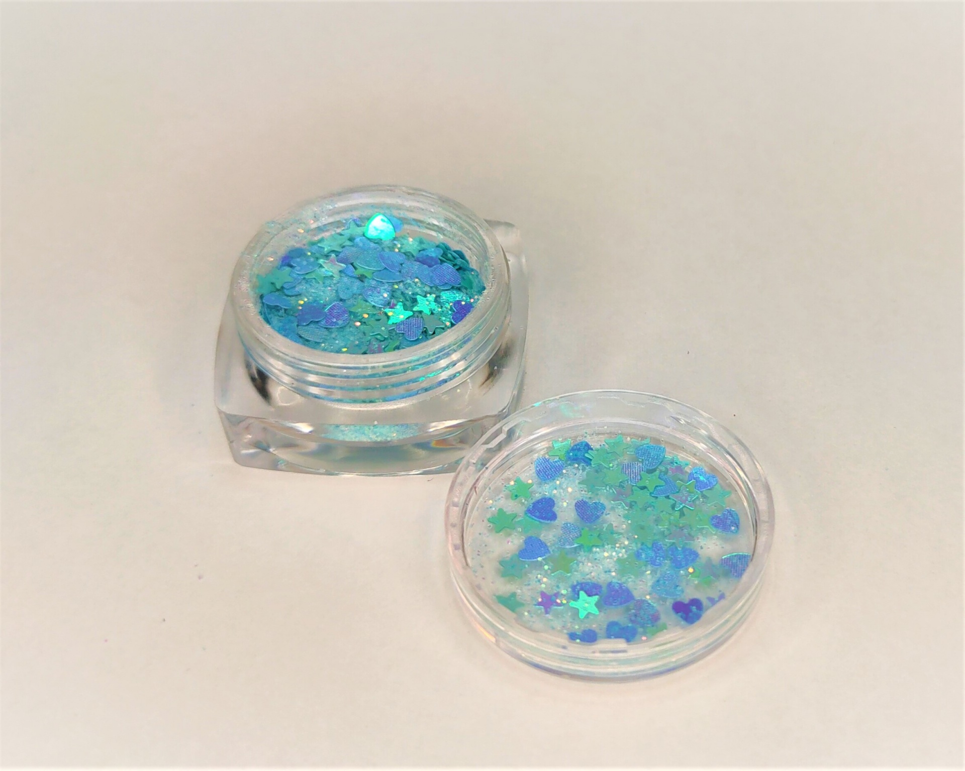 Dois pequenos potes transparentes com glitter colorido em formas variadas sobre fundo branco.