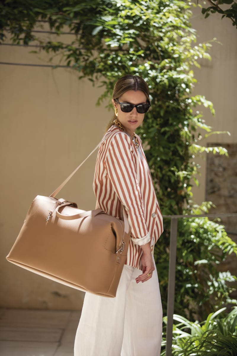 Mulher com blusa riscada, calças brancas e mala castanha em pele ao ar livre