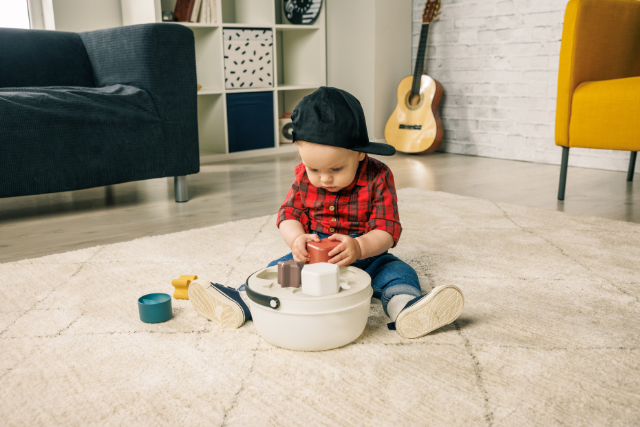 Bebé a brincar com brinquedos de madeira num tapete bege em sala com sofá cinzento e cadeira amarela
