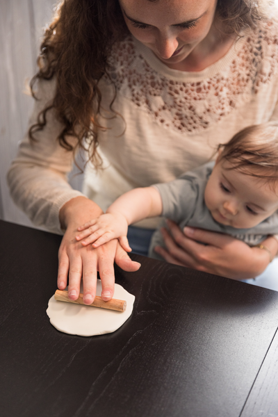 Mulher com camisola bege e bebé a esticar massa branca numa mesa preta