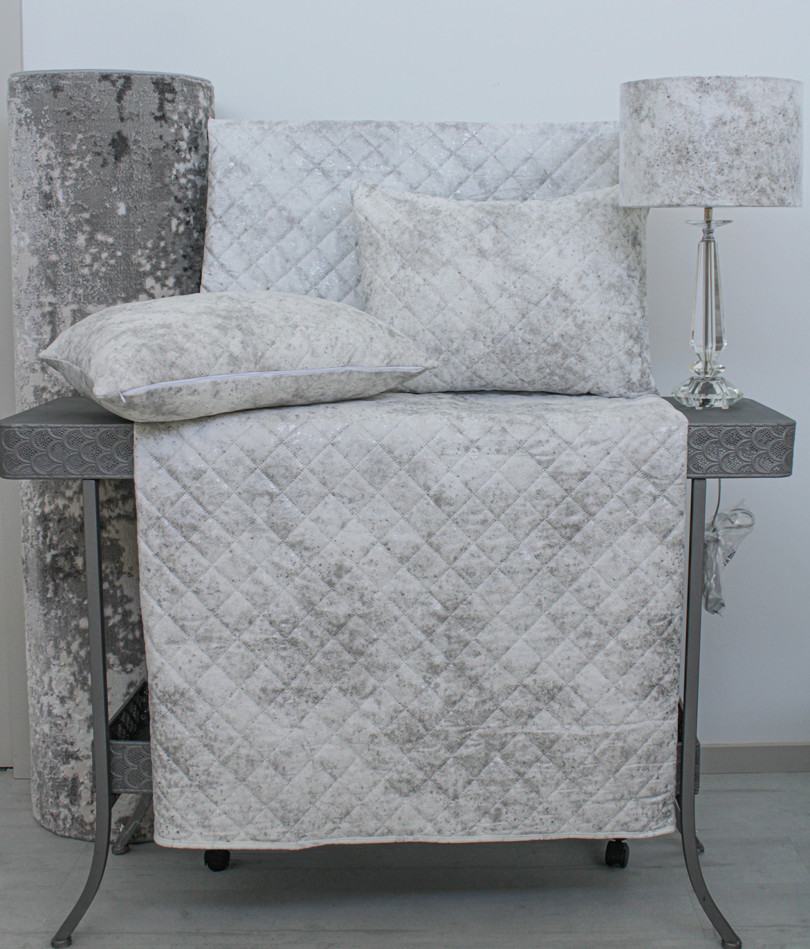 Conjunto de têxteis cinza claro com padrão de losangos e almofadas sobre mesa cinza com lâmpada.