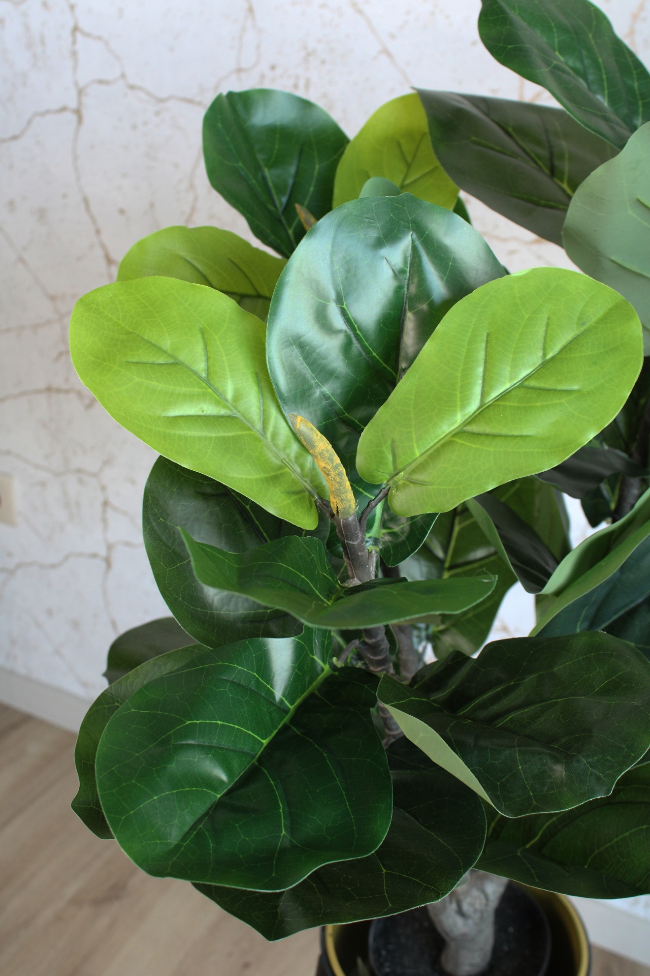 Planta artificial com folhas verdes em vaso preto com borda dourada junto a parede bege