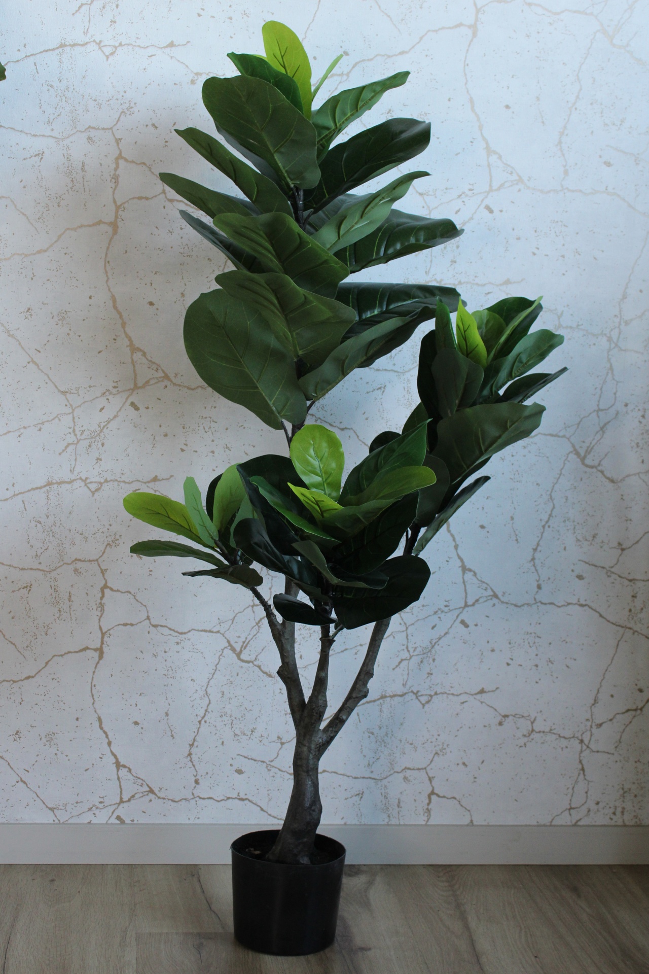 Planta artificial verde em vaso preto num piso cinzento claro com parede branca e padrão rachado