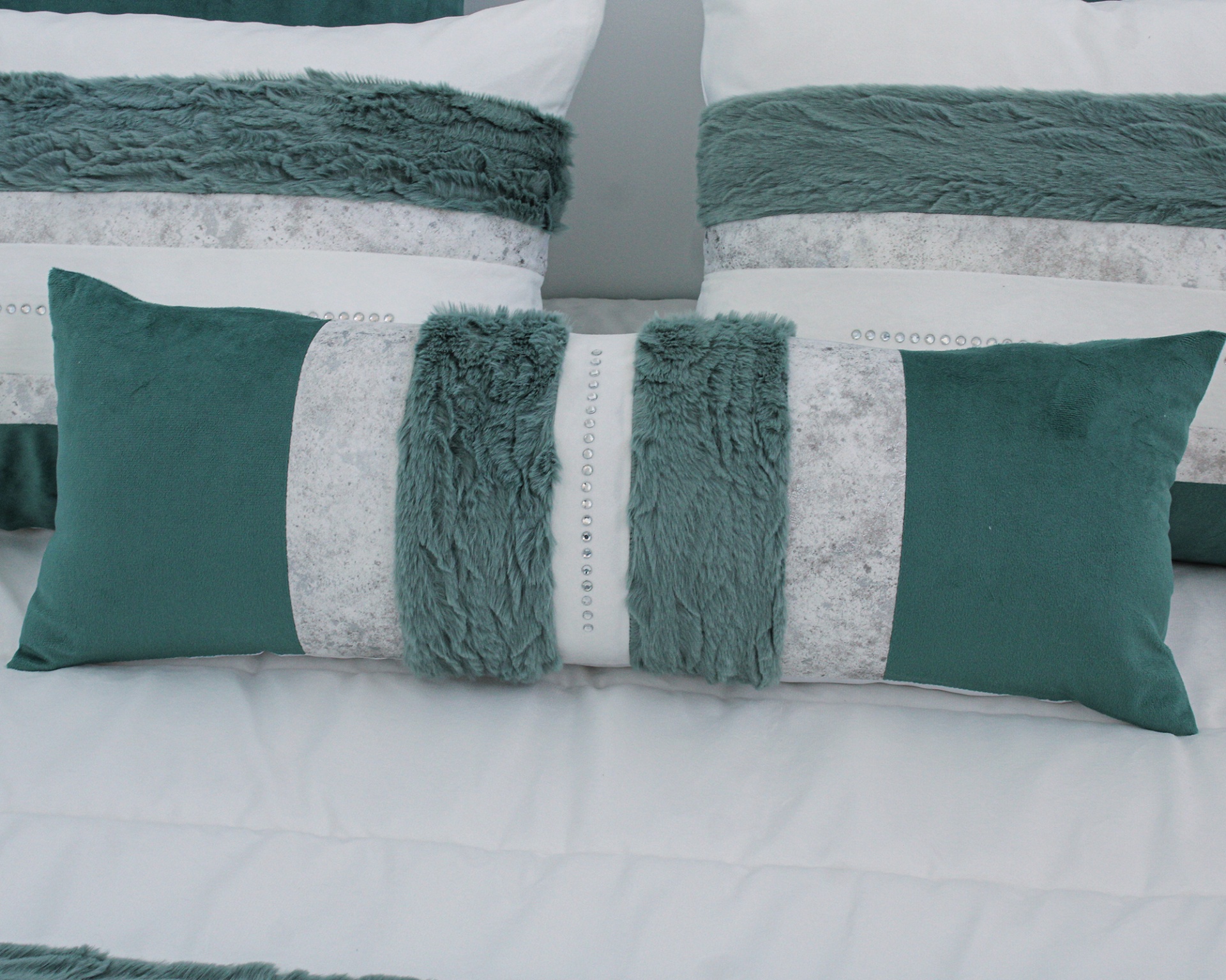 Três almofadas decorativas verde água e cinza com texturas felpudas e tachas metálicas numa cama