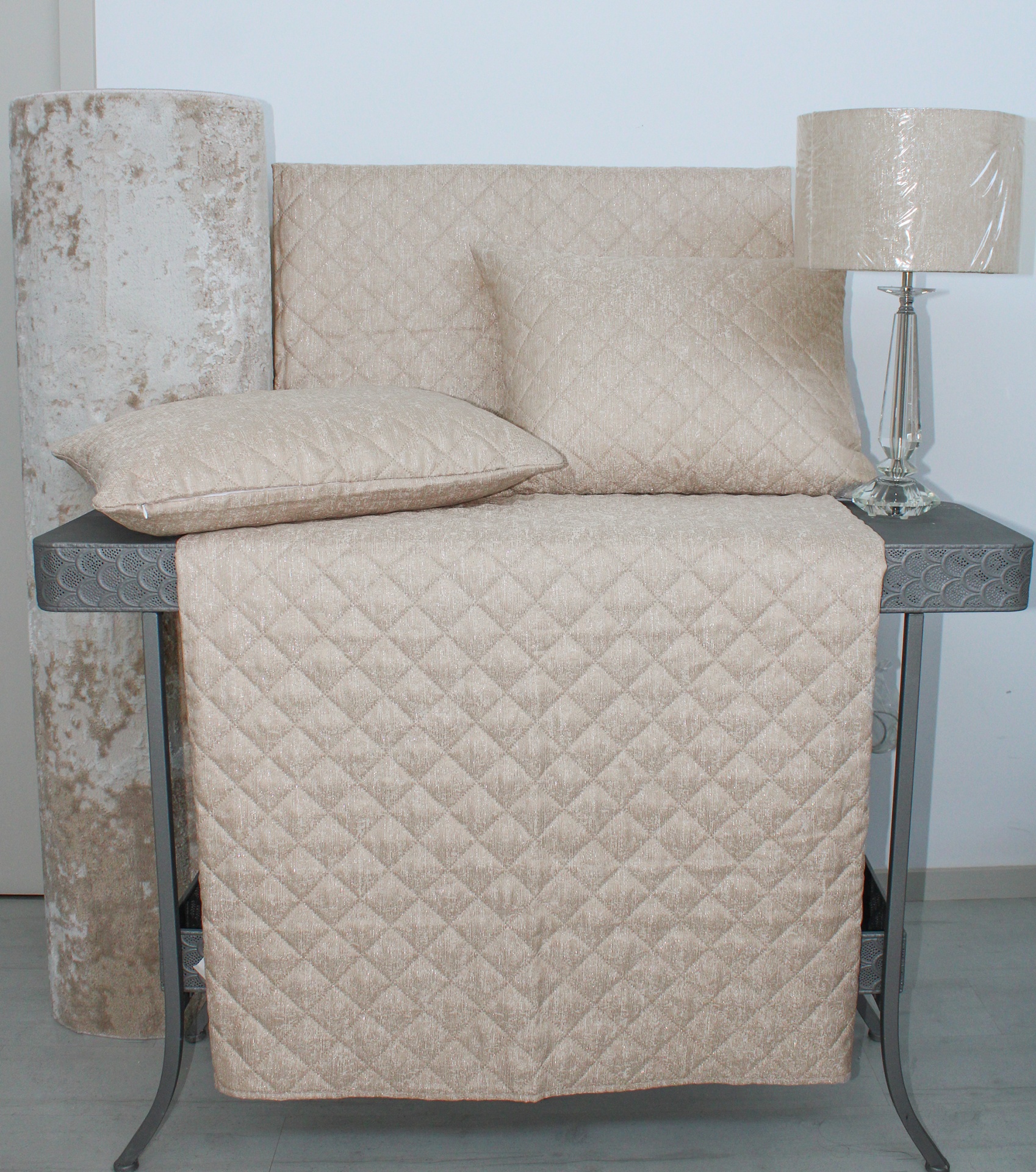 Conjunto de almofadas e manta bege acolchoadas com padrão de quadrados, rolo de tecido e lâmpada de mesa