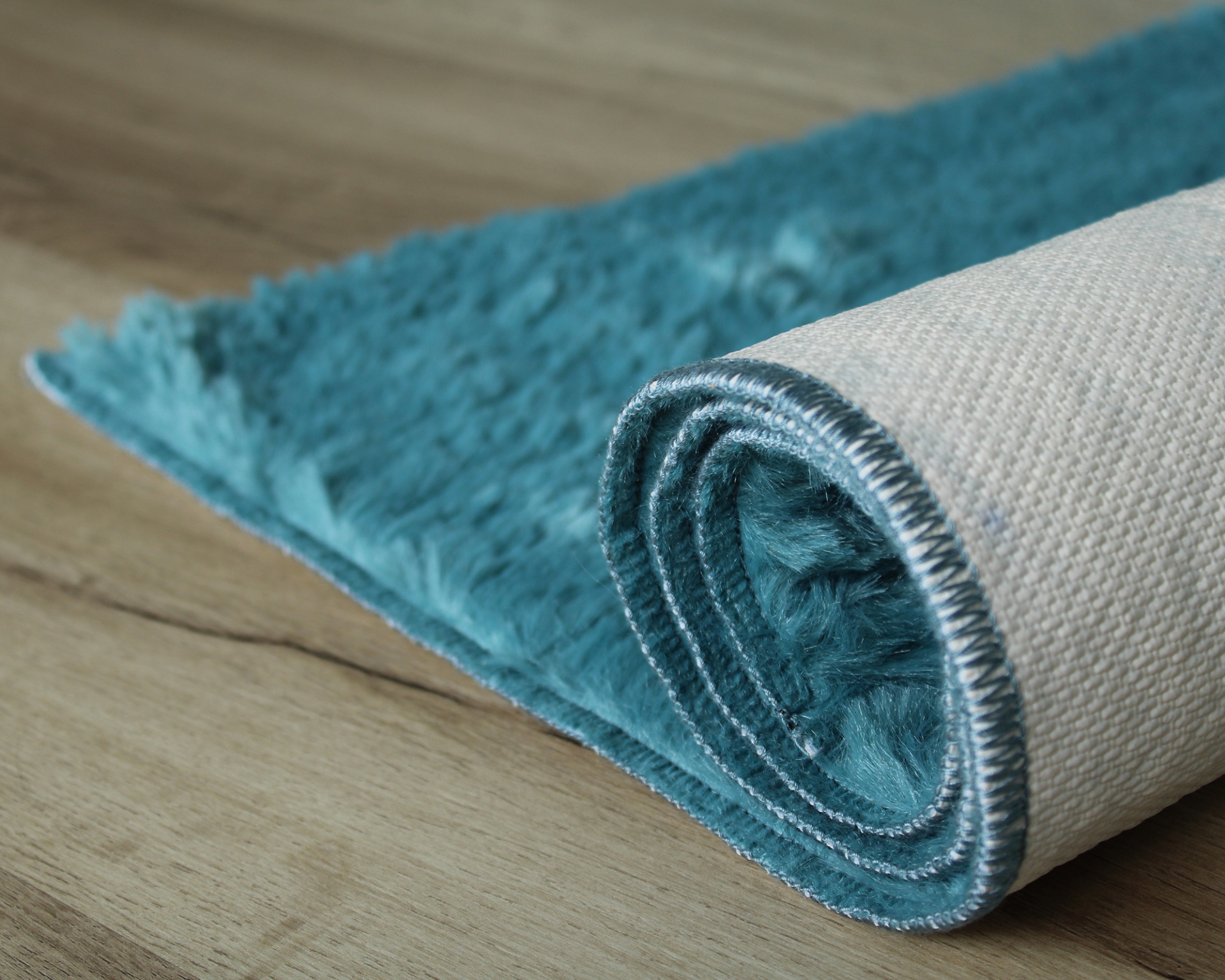 Tapete azul turquesa felpudo enrolado mostrando verso em tecido bege sobre chão de madeira clara