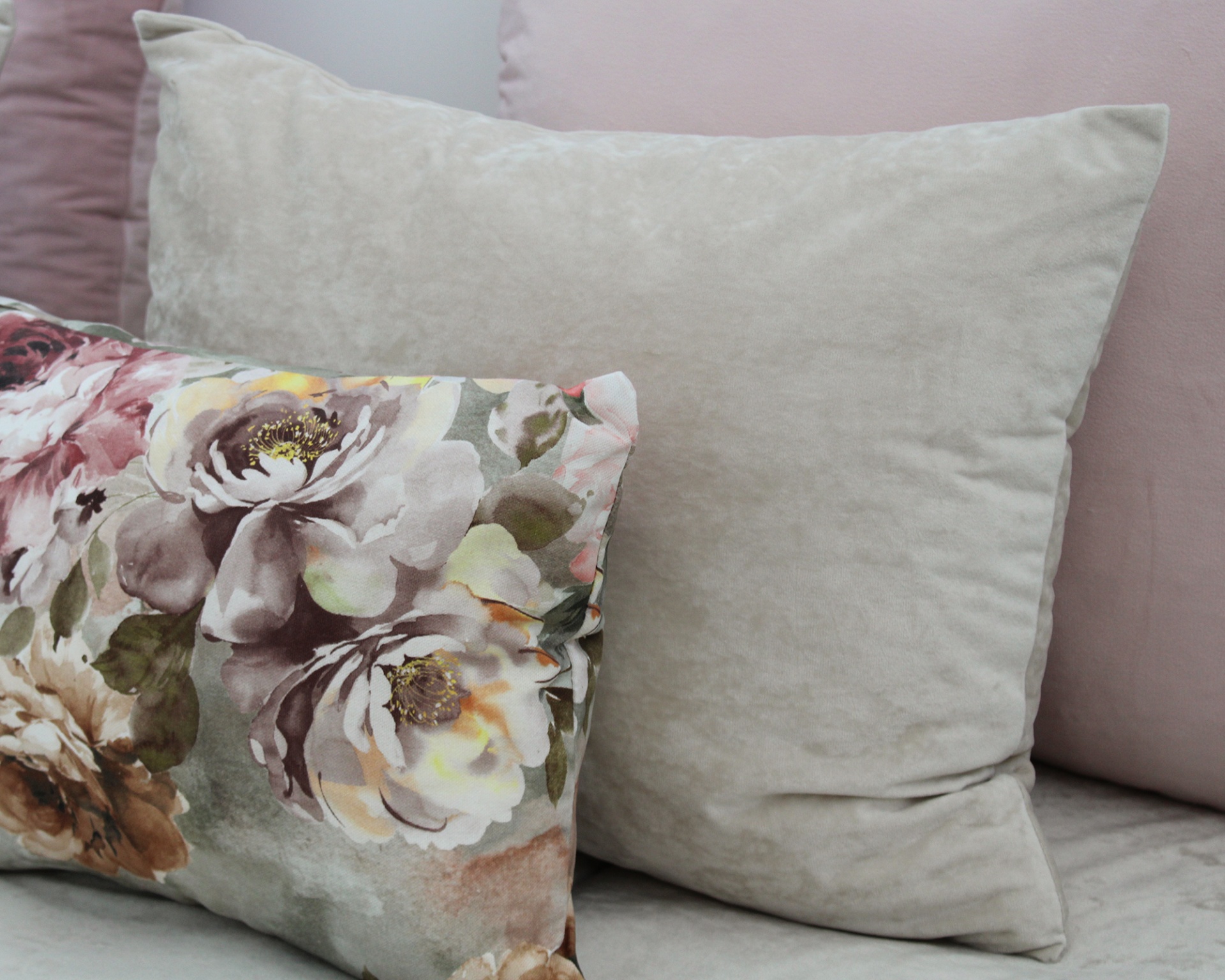 Três almofadas decorativas em tons pastel, incluindo uma lisa cinza, uma lisa rosa e uma com padrão floral colorido.