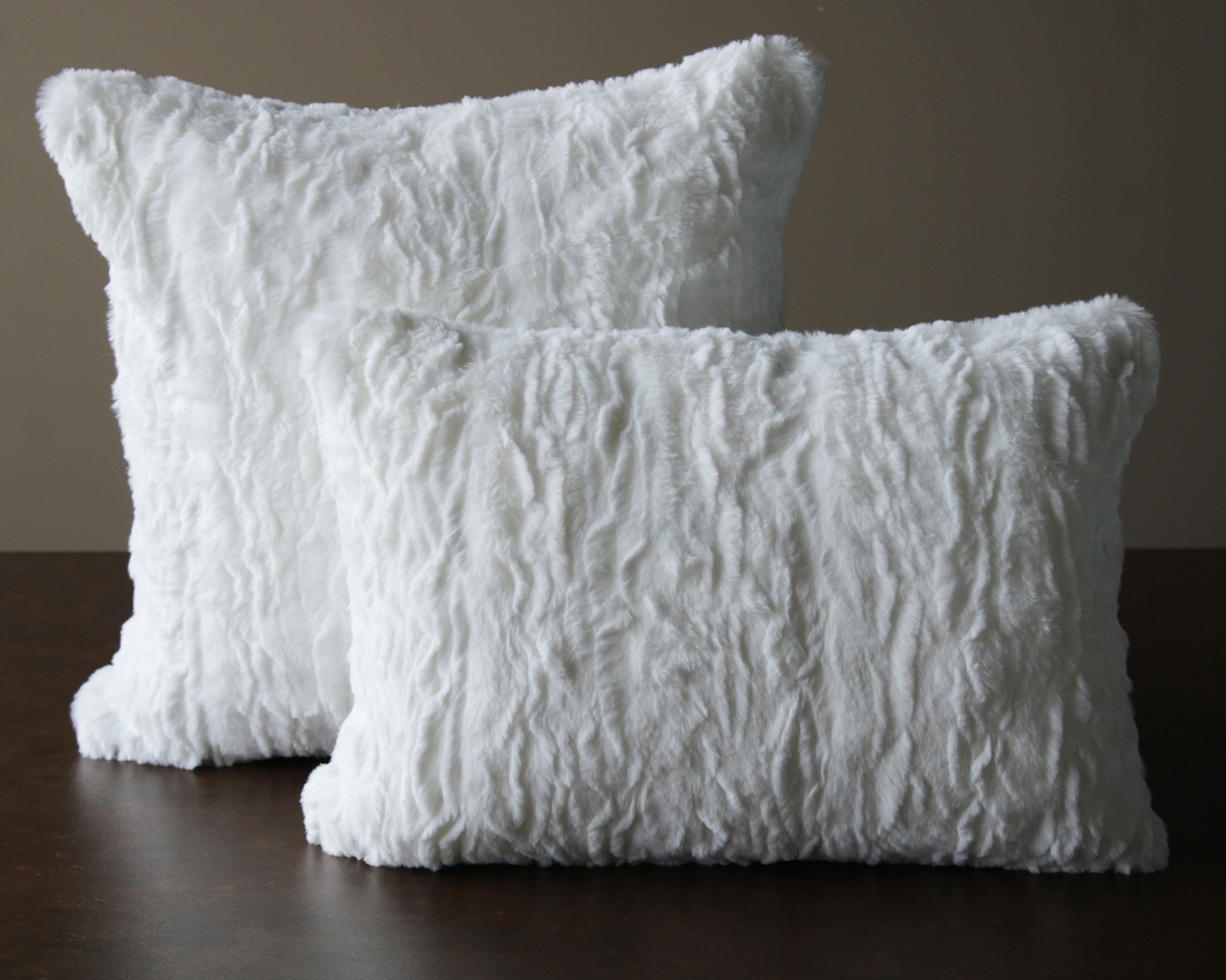 Duas almofadas brancas felpudas, uma quadrada e outra retangular, sobre superfície castanha.