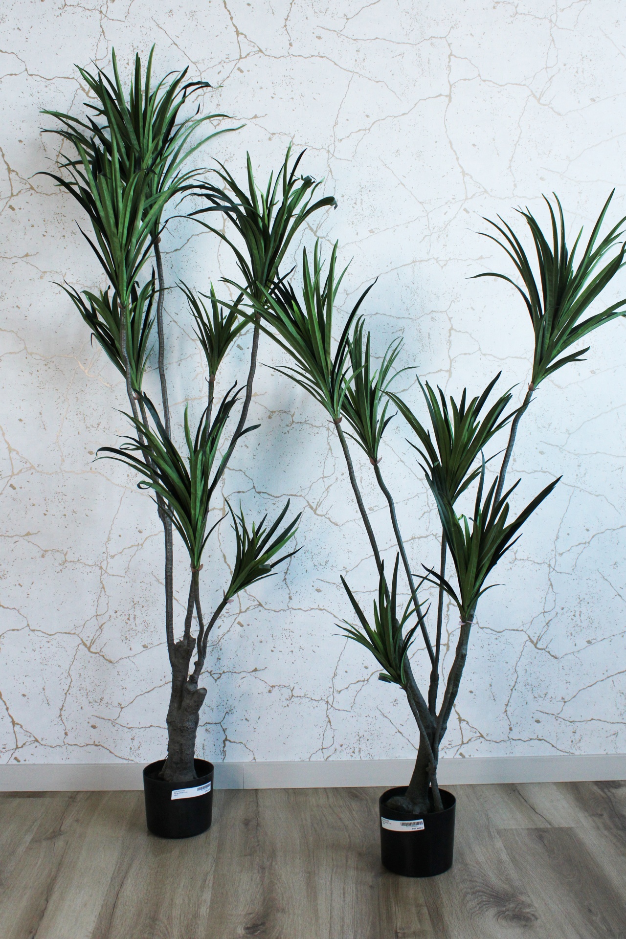 Duas plantas artificiais em vasos pretos junto a parede branca texturada