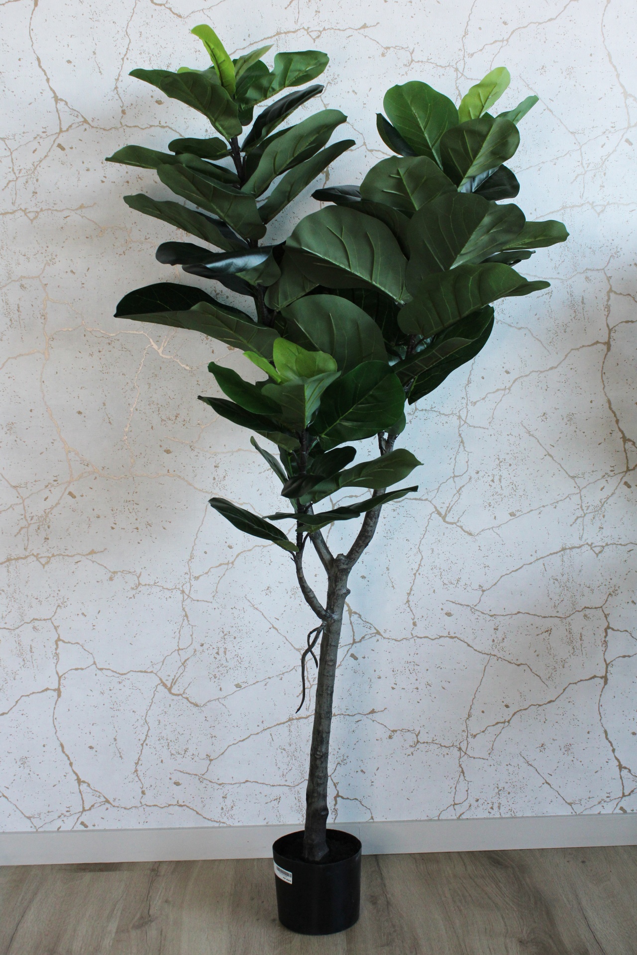 Planta artificial com folhas verdes largas em vaso preto junto a parede branca riscada