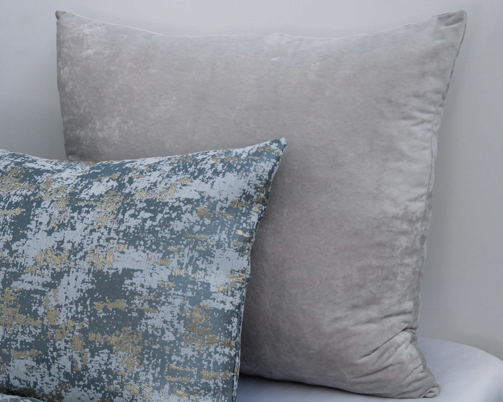 Dois almofadas decorativas, uma cinzenta aveludada e outra com padrão azul, cinza e dourado.