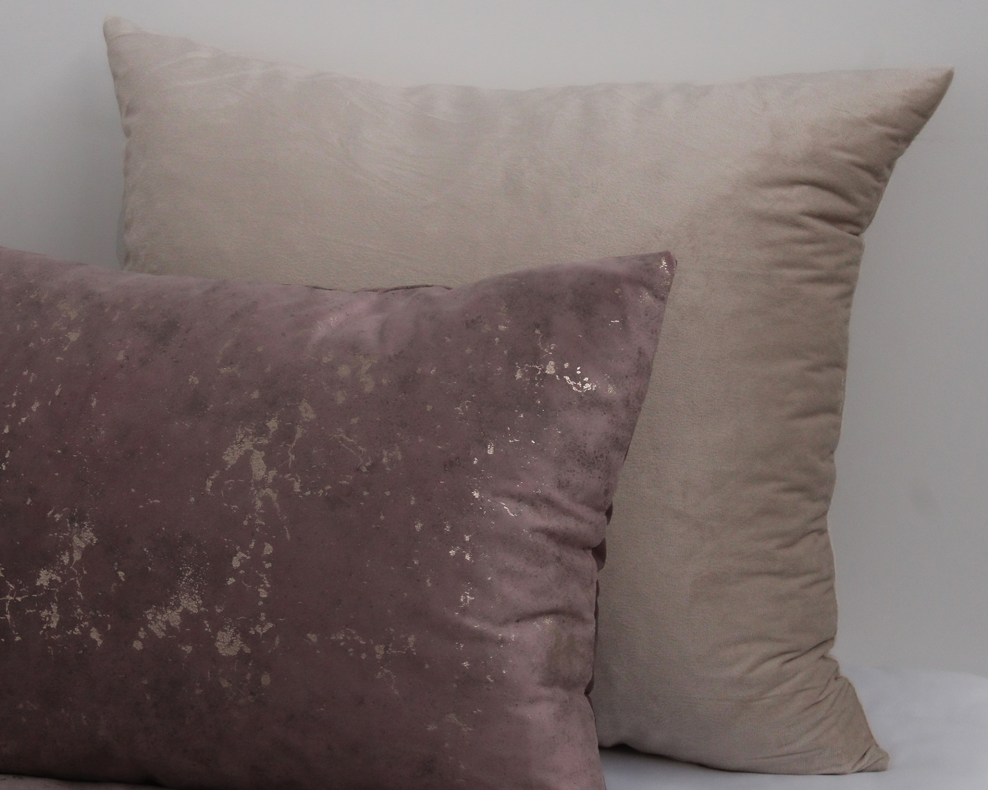 Duas almofadas retangulares, uma bege lisa e outra roxa com padrão metálico.