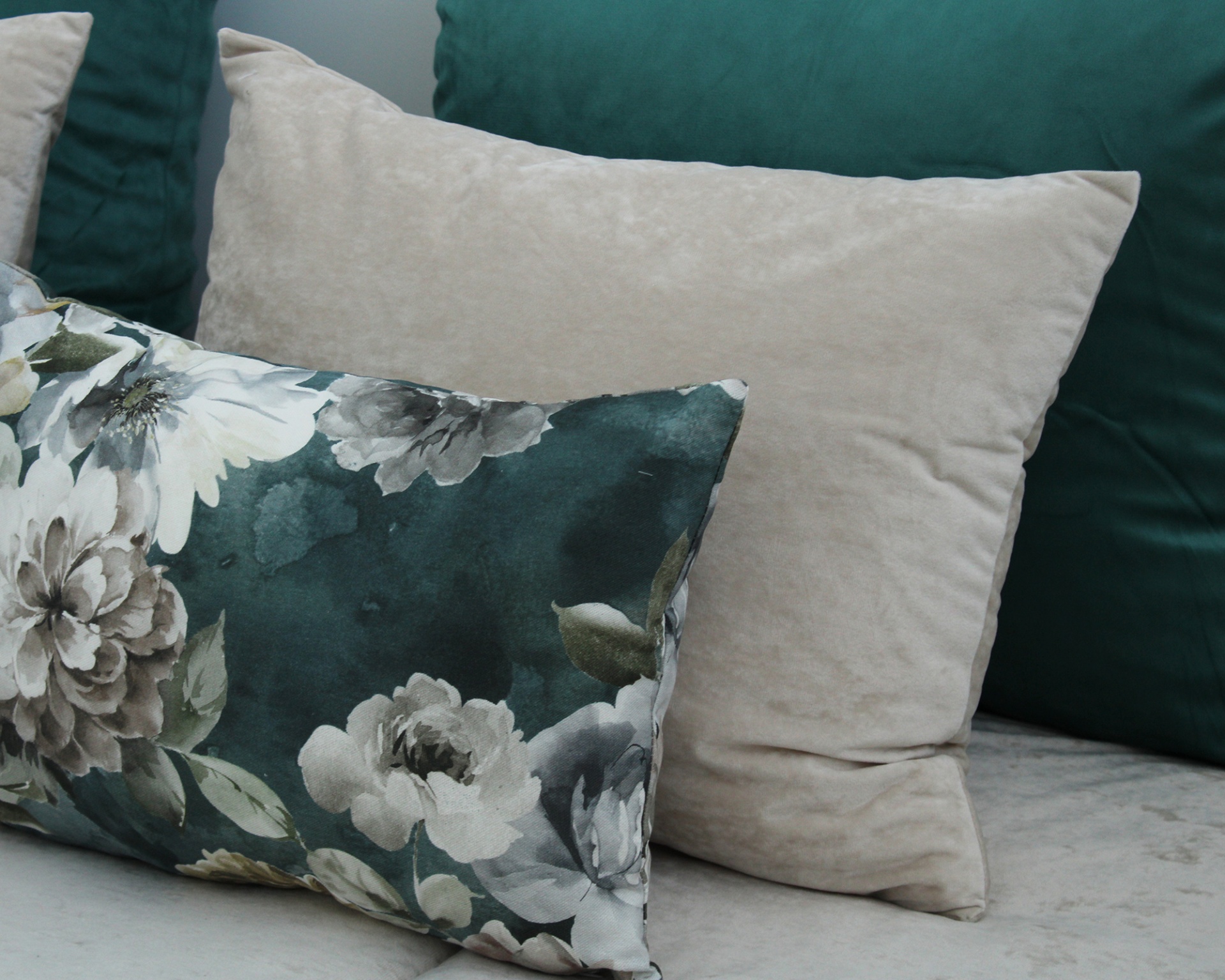Três almofadas em tecido macio, uma bege lisa, uma verde escura lisa e uma floral em tons de verde e bege