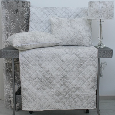 Conjunto de têxteis cinza claro com padrão de losangos e almofadas sobre mesa cinza com lâmpada.