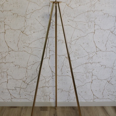 Candeeiro de pé metálico dourado com três pernas e suporte para lâmpada num chão em madeira e parede branca com padrão rachado