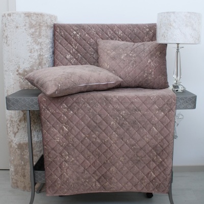 Colcha castanha acolchoada com almofadas, cortinas bege enrolladas, mesa cinza e luminária com abajur branco