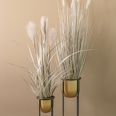 Dois vasos dourados com plantas artificiais em suportes pretos, chão de madeira e parede bege.