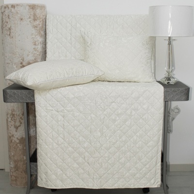 Cobertura e almofada acolchoadas em tecido acetinado branco com padrão de losangos sobre mesa escura, com rolo de tecido bege e candeeiro branco ao lado