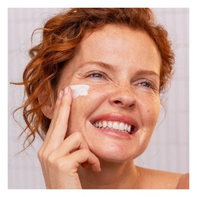 Mulher aplica creme no rosto com sorriso e cabelo ruivo encaracolado