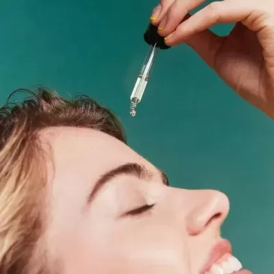 Pessoa aplicando óleo facial com conta-gotas, fundo azul esverdeado
