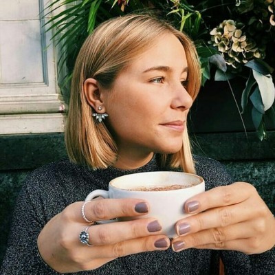 Mulher loira com camisola cinza segurando uma caneca branca, usando anéis e brincos prateados.
