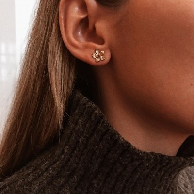 Brinco pequeno dourado em forma de flor com pedras claras no centro na orelha de uma pessoa