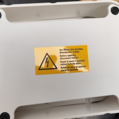 Parte inferior de dispositivo branco com etiqueta amarela de aviso elétrico e pés de borracha pretos