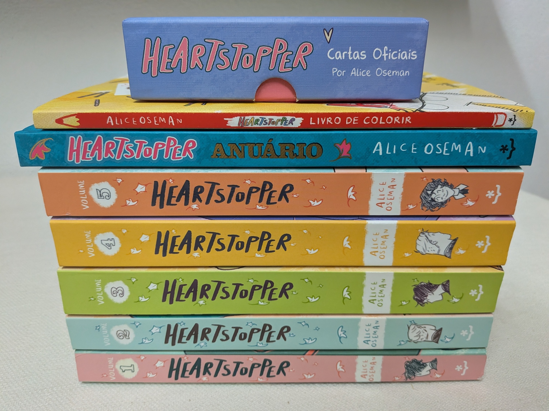 Alice Oseman - FULL PACK Heartstopper (5 volumes + Livro de Colorir + Anuário + Baralho de Cartas) Livros da coleção Heartstopper empilhados por Alice Oseman com diferentes capas coloridas.
